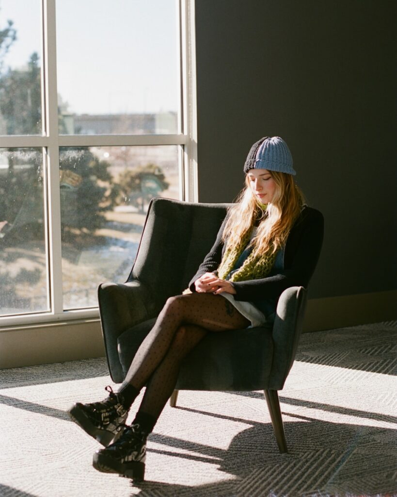 Young woman in hat sits in armchair by window.